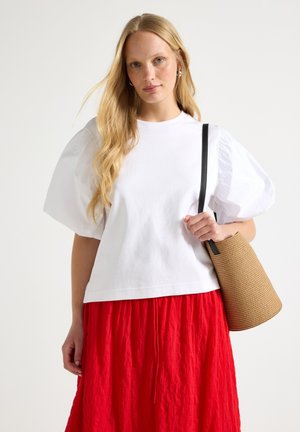 Donna con lunghi capelli biondi indossa una maglia bianca con maniche a sbuffo e una gonna rossa, tenendo una borsa a secchiello marrone intrecciata con tracolla nera sulla spalla.