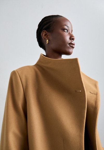 Camel wool coat with high collar, minimal design, and subtle stitching details. Model wears gold hoop earrings, showcasing a clean texture.