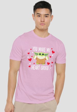 Mann trägt hellrosa T-Shirt mit Cartoon-Grogu, umgeben von roten Herzen und dem Text „Du machst mein Herz Grogu“ vorne.