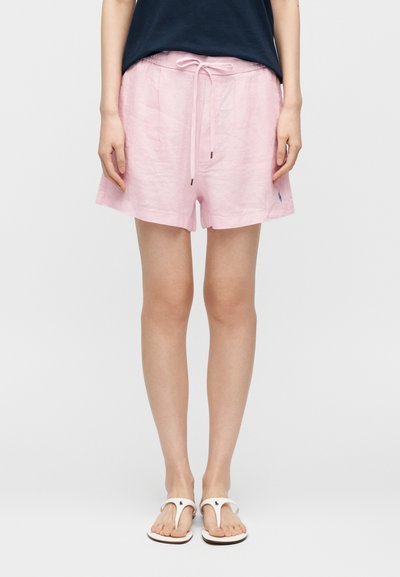 Άτομο που φορά ανοιχτόροζ σορτς με κορδόνι, μπλε τοπ και λευκές σαγιονάρες, στέκεται σε απλό λευκό φόντο.