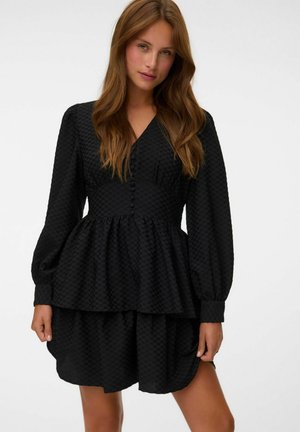 Femme aux longs cheveux bruns portant une robe noire texturée à manches longues avec une taille boutonnée et une jupe évasée sur un fond uni.