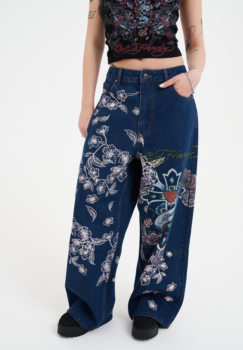 Vaqueros de mezclilla ligeros de pierna ancha con bordados florales en blanco, que presentan gráficos de estilo tatuaje coloridos y un ajuste relajado. Acentos negros en los zapatos.