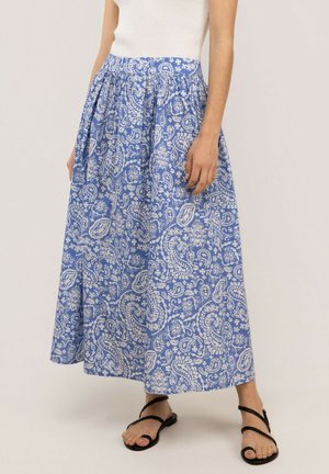 Mujer con falda larga con patrón paisley en azul y blanco, top blanco sin mangas y sandalias negras con tiras trenzadas, de pie.