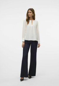 Blusa bianca a maniche lunghe con scollatura a chiave, abbinata a jeans blu scuro a gamba larga. Il tessuto presenta una leggera texture e una vestibilità strutturata.