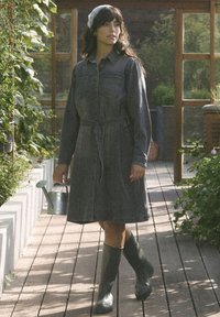 Robe en jean, gris foncé, à manches longues, devant boutonné, taille ceinturée, avec deux poches poitrine ; portant des bottes en caoutchouc vertes, tenant un arrosoir.