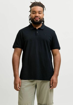 Szakállas, raszta hajú férfi fekete galléros pólót és bézs nadrágot visel, egy egyszínű világos háttér előtt áll.