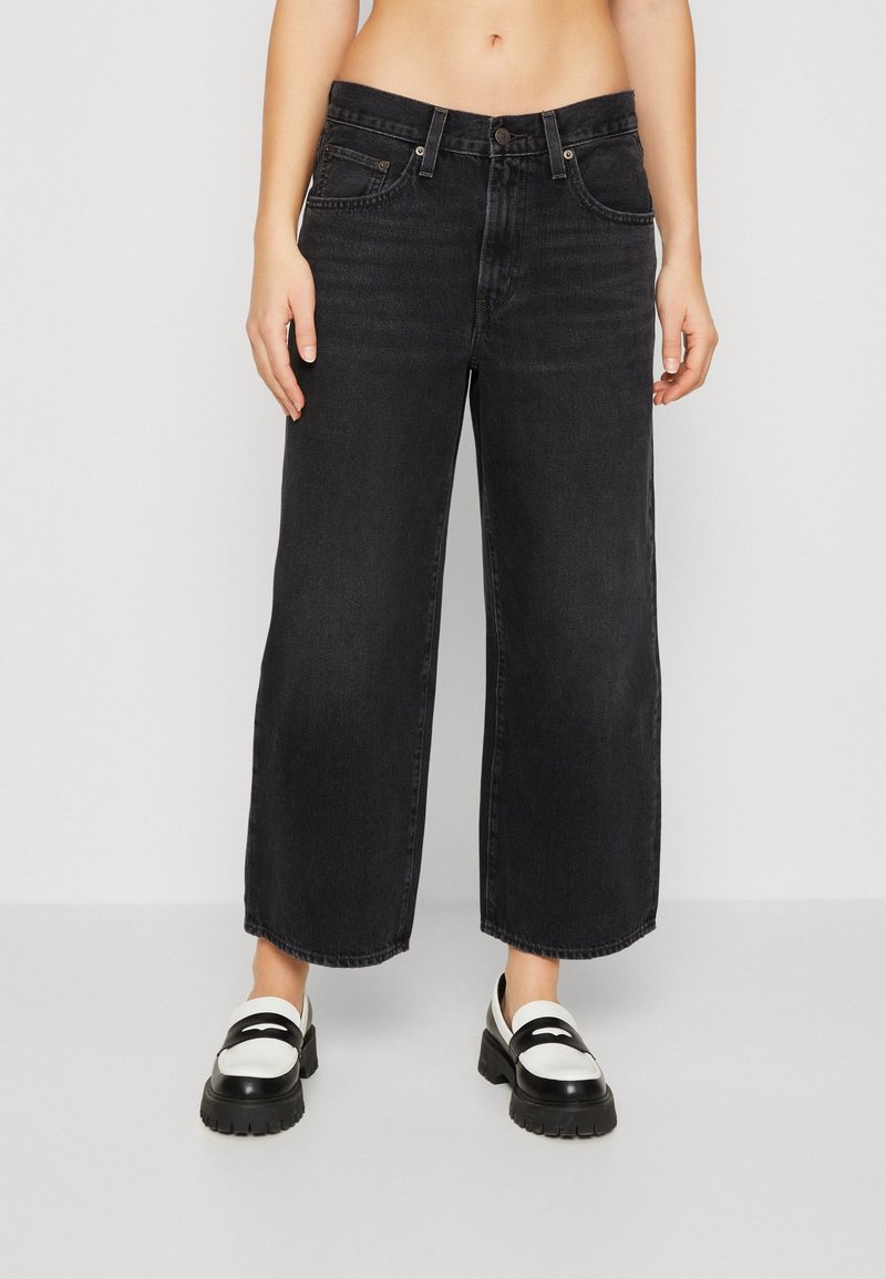 Personne portant un jean noir taille haute à jambes larges, coupé au-dessus des chevilles, avec des mocassins épais blancs et noirs sur un fond uni.