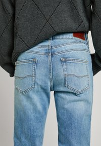 Ljusblå denimjeans med rak skärning, synliga bakfickor och orange sömmar; matchat med en mörkgrå stickad tröja.