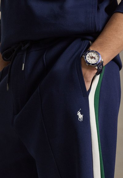 Personne portant un pantalon bleu marine avec des bandes latérales blanches et vertes, une main dans la poche, et une montre-bracelet bleue et argentée au poignet.