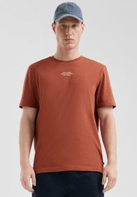 Koperkleurig katoenen T-shirt met een ronde hals, korte mouwen en witte tekstprint op de borst. Gecombineerd met een lichtblauwe pet.