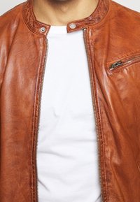 Brun läderjacka med en slimmad design, med dragkedja, tryckknappskrage och en ficka med dragkedja på bröstet. Bärs över en vit t-shirt.