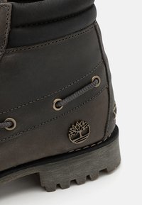 Donkergrijze leren laars met een ronde neus, metalen oogjes, gestructureerde stiksels en een ronde embleem met een boom aan de zijkant.
