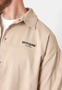 Chaqueta beige de botones con cuello puntiagudo, hecha de tejido texturizado, con el logotipo bordado en negro "SIXTHJUNE PARIS" en el lado izquierdo.