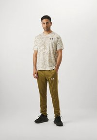 Lichtbeige camouflage T-shirt met korte mouwen en zwart logo. Olijfgroene joggingbroek met wit logo, gecombineerd met zwarte sneakers.
