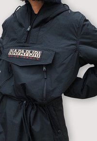 Chaqueta tipo pulóver negra con capucha, que cuenta con un bolsillo delantero, cierre de cremallera y cintura ajustable con cordón. Logo mostrado en el bolsillo.