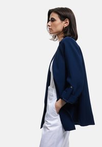Blazer navy con vestibilità ampia, scollatura rotonda e maniche arrotolate, abbinato a una maglietta bianca e pantaloni dal design minimalista.