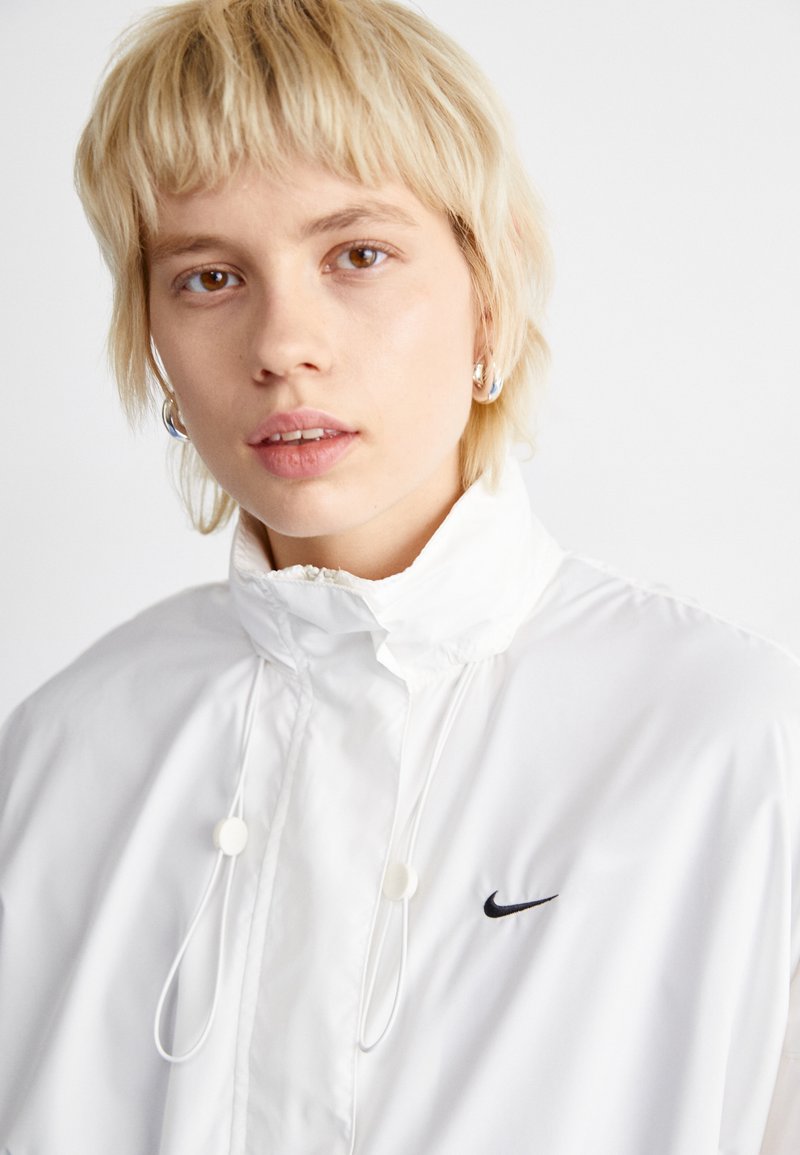 Witte lichtgewicht jas met hoge kraag, voorzien van een zwart Nike-logo en verstelbare trekkoorden. Soepele textuur en minimalistisch ontwerp.