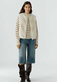 Nepbont gilet in crème over een gestreepte trui in beige en bruin. Gecombineerd met wijdvallende blauwe jeans en knie-hoge bruine laarzen.