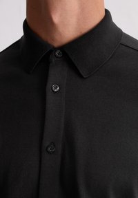 Schwarzes Poloshirt mit Kragen, ausgestattet mit einer Knopfleiste mit schwarzen Knöpfen, aus Strickstoff gefertigt. Glatte Textur mit taillierter Passform.