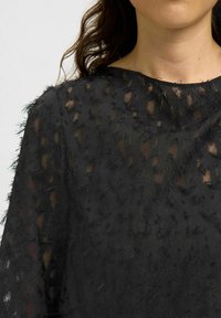 Top nero trasparente in tessuto testurizzato, con motivi irregolari e bordi sfrangiati. Collo rotondo, design leggero adatto per essere indossato a strati.