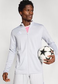 Αθλητικό μπουφάν σε ανοιχτό γκρι χρώμα με φερμουάρ μπροστά, ψηλό γιακά και λογότυπα. Το μοντέλο κρατά μια ποδοσφαιρική μπάλα, φοράει ροζ φανελάκι. Ομαλή υφή.