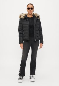 Casaco puffer preto com capuz forrado de pelo, combinado com uma blusa preta justa e jeans escuros com detalhes em botão, finalizado com ténis pretos.