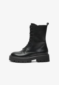 Svart lær lace-up militærstøvel med tykk såle, ankelhøy skaft og dragløkke bak på hvit bakgrunn.