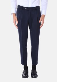 Pantaloni tailored navy con pieghe frontali, passanti per cintura e chiusura con bottone. Texture liscia, lunghezza cropped, abbinati a scarpe nere.