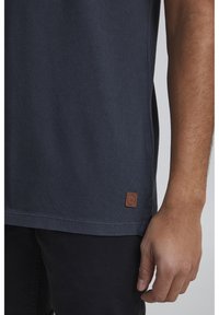 Námořnická košile s krátkým rukávem z texturované tkaniny s hnědou koženou visačkou na lemu, v kombinaci s tmavými kalhotami.