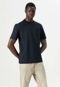 Camiseta de algodón azul marino con cuello redondo, mangas cortas y un bolsillo en el pecho. Presenta costuras en contraste a lo largo de las costuras.