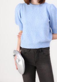 En person som bär en ljusblå kort puffärmad tröja och svarta jeans, håller en silverfärgad mjuk clutchväska med ena handen.