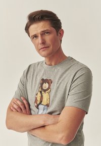 Szürke pamut póló, amelyen egy sárga kapucnis pulcsit viselő medve illusztrációja látható, aki gitárt tart. A mintázat részletes varrással és díszítésekkel van ellátva.