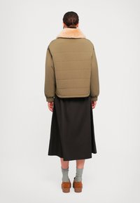 Donna con i capelli legati indossante un giacca imbottita verde oliva con colletto di pelliccia beige, gonna midi nera, calze grigie e scarpe marroni, di spalle.