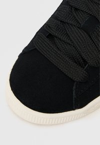 Juodas zomšinės sportbačiai su pintomis juodomis raištelėmis, apvaliu kančių ir balta gumine apsole. Viršus turi matinį paviršių ir subtilų siuvinėjimo detalių.