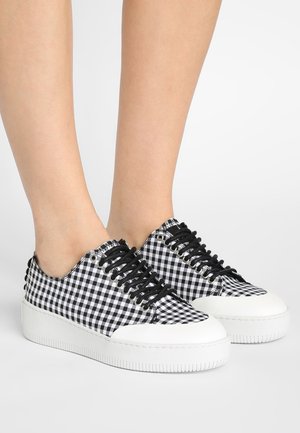 Stopy w czarno-białych niskich sneakersach w kratkę gingham, z czarnymi sznurowadłami i grubymi białymi gumowymi podeszwami na białym tle.
