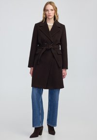 Cappotto in misto lana marrone con design doppiopetto, cintura in vita, grandi collo e tasche laterali, abbinato a jeans a gamba larga blu e tacchi neri.