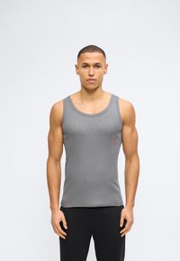 Μυώδης άνδρας που φοράει μια εφαρμοστή γκρι αμάνικη φανέλα με ένα μικρό κόκκινο λογότυπο «Hugo» στο αριστερό στήθος, στέκεται μπροστά σε απλό λευκό φόντο.