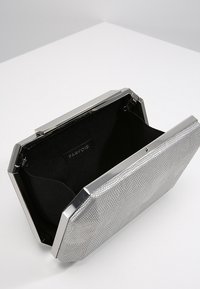 Silver textured rectangular clutch bag with metallic frame, open to show black fabric interior and brand label inside.