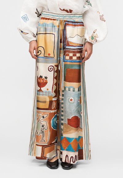 Pantalon à jambes larges avec des motifs abstraits et géométriques colorés dans des tons terre, associé à un chemisier blanc à manches longues avec des broderies sur les manches.