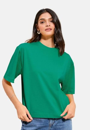 Femme aux cheveux foncés mi-longs portant un T-shirt vert à manches courtes et un jean bleu, posant avec une main sur la hanche devant un fond blanc.