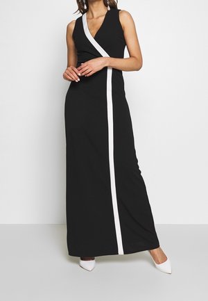 Donna che indossa un vestito nero senza maniche con una striscia diagonale bianca e scarpe bianche a punta, in piedi davanti a uno sfondo semplice.