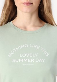 T-shirt en coton vert clair à manches courtes. Le texte en blanc indique "RIEN DE TEL QUE CET ÉTÉ MAGNIFIQUE" dans un design incurvé.