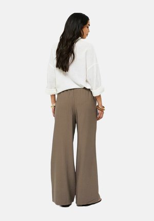 Vrouw met lang donker haar, gekleed in een witte blouse met lange mouwen en brede bruine broekspijpen, gezien van achteren, staande tegen een effen achtergrond.