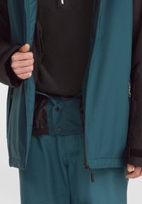 Chaqueta exterior en color teal con detalles en negro, que presenta un cierre frontal con cremallera, puños ajustables y un cierre a presión en la cintura. El interior muestra un diseño en capas.