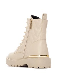 Bottines beige en matériau synthétique matelassé, avec une fermeture éclair sur le côté, un accent doré sur le talon et une semelle épaisse texturée.
