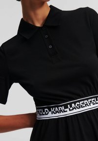 Svart poloklänning med korta ärmar, med en knappad krage och ett elastiskt midjeband som visar "KARL LAGERFELD" i vit text.