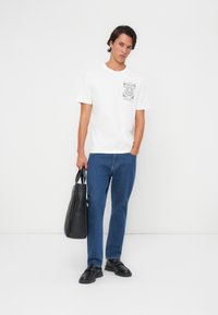 T-shirt en coton blanc avec un graphisme noir sur la poitrine, associée à un jean en denim bleu et des mocassins noirs, tenant un sac fourre-tout noir.