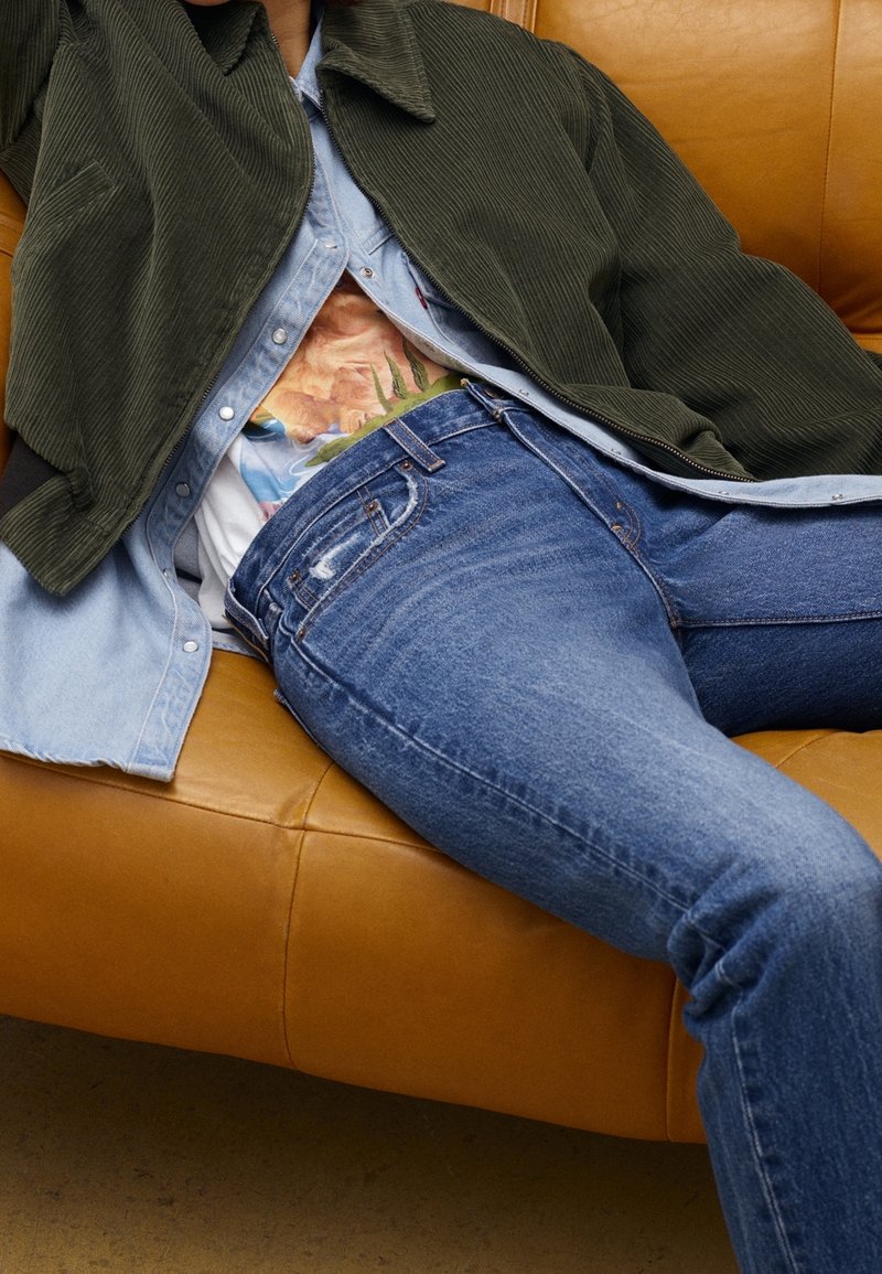 Personne allongée sur un canapé en cuir marron portant un jean bleu, une chemise à motifs, une chemise bleu clair boutonnée et une veste en velours côtelé vert foncé.