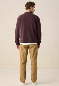 Mann steht mit dem Rücken zur Kamera, trägt ein dunkellila Langarmoberteil, beigefarbene Cordhosen, weiße Sneakers und ein weißes Hemd darunter.