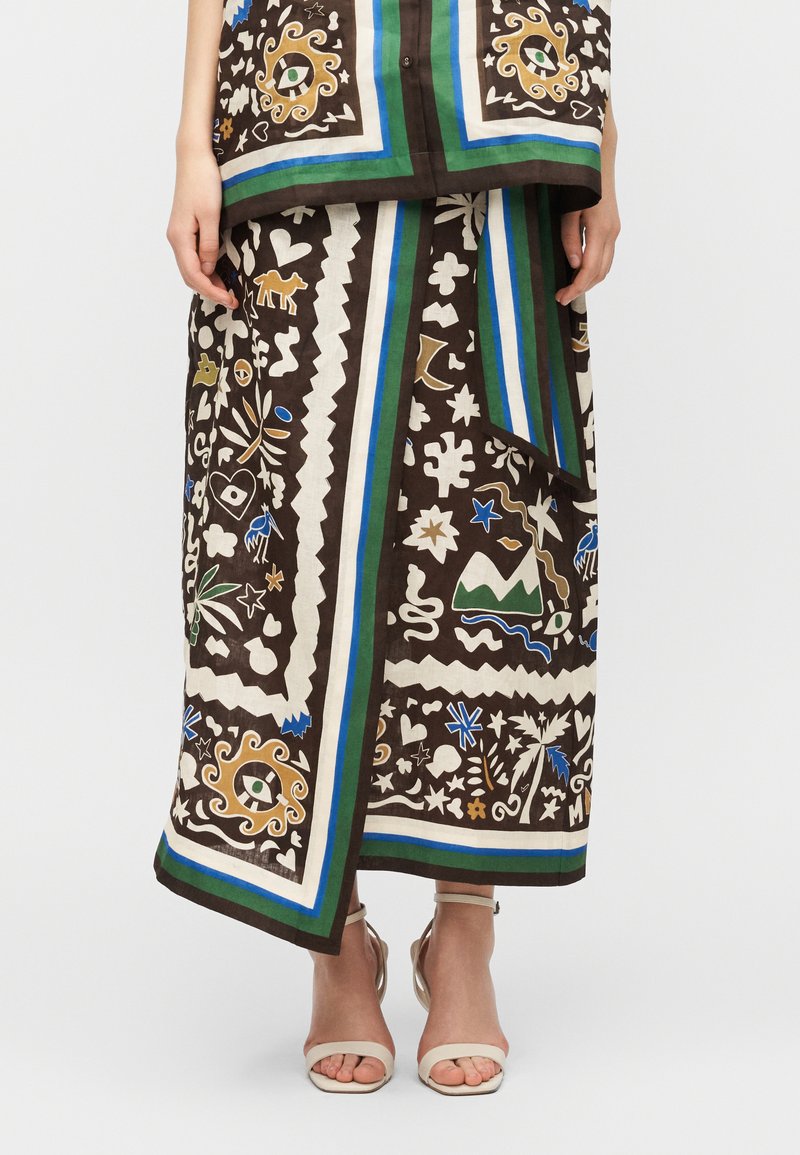 Γυναίκα που φοράει καφέ περιτυλιγμένη φούστα κα ι τοπ με πολύχρωμα γεωμετρικά και εμπνευσμένα από τη φύση σχέδια, καθώς και λευκά ψηλοτάκουνα πέδιλα με ανοιχτές μύτες, σε λευκό φόντο.